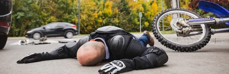 A motorcyclist in protective gear lies injured on the road next to a fallen motorcycle.